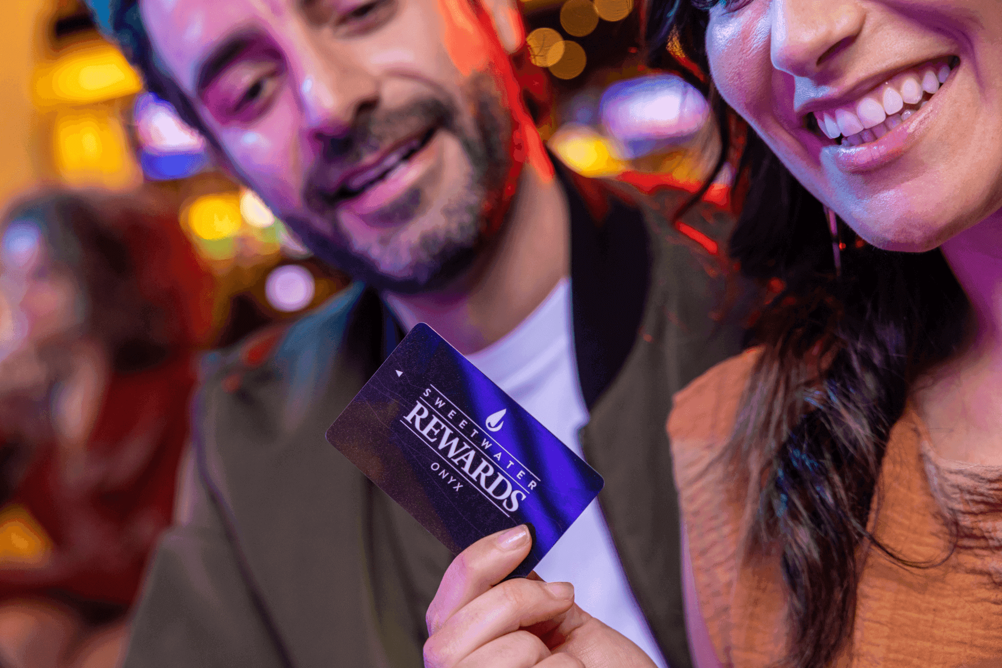 A smiling woman and a man displaying a Sweetwater Rewards Onyx membership card at a lively venue, symbolizing exclusive customer benefits.